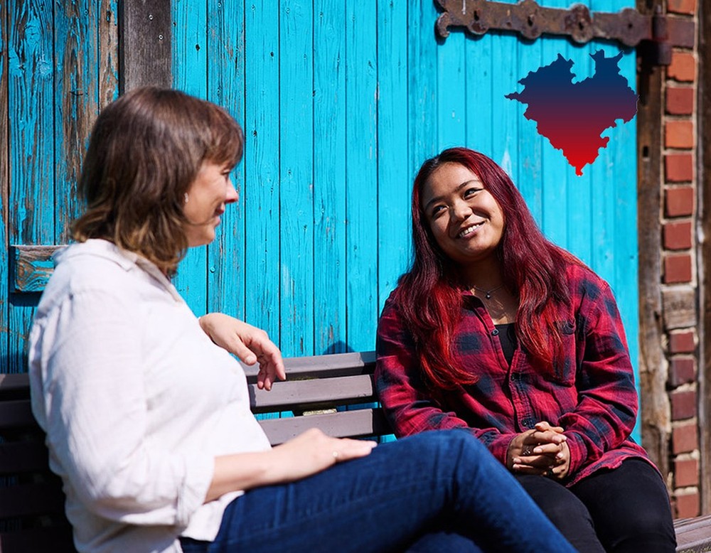 Zwei Frauen unterhalten sich auf einer Bank vor einer blauen Holzwand. Daneben ein illustriertes Westfalenherz